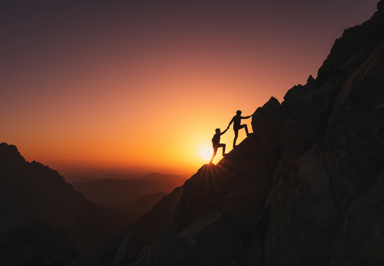 Two climbers helping one another reach a mountain summit at sunset